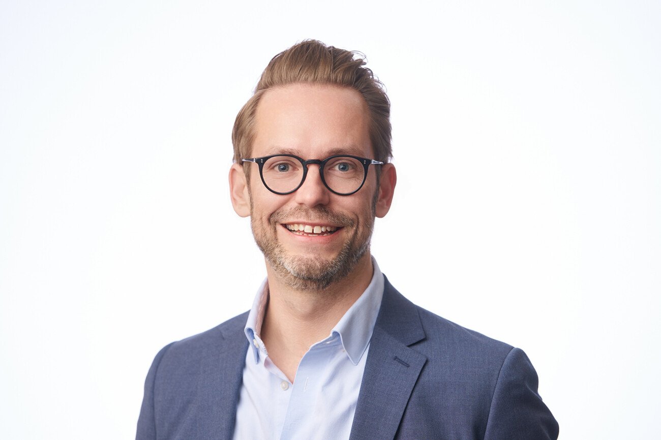 Smiling man with glasses and short hair, in a blue suit and light blue shirt, captured expertly by a corporate headshot photographer against a plain white background. James Gifford-Mead Photography - Event Photographer London