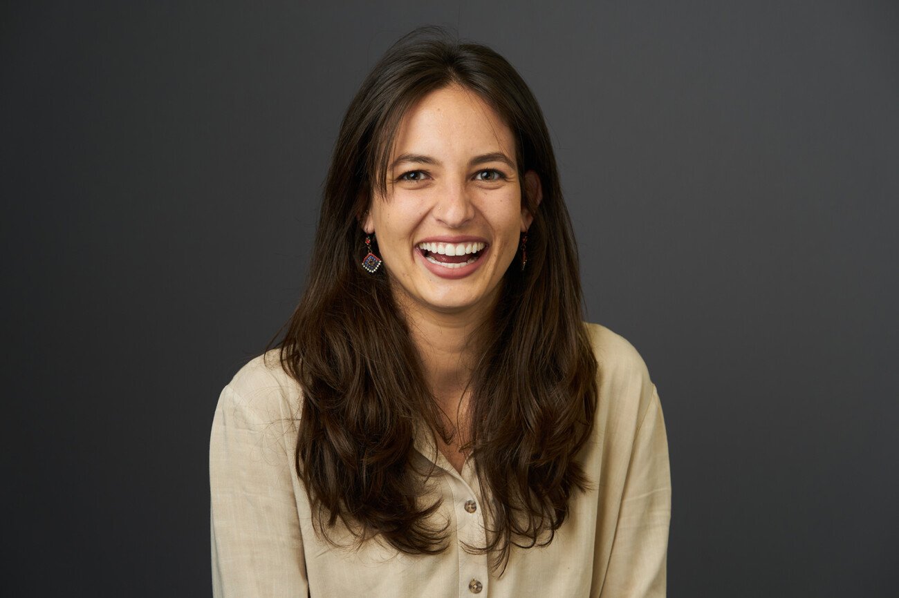 A woman with long brown hair smiles confidently while looking forward, showcasing the expertise of a corporate headshot photographer. She is wearing a beige blouse and colorful earrings, standing against a plain dark background. James Gifford-Mead Photography - Event Photographer London