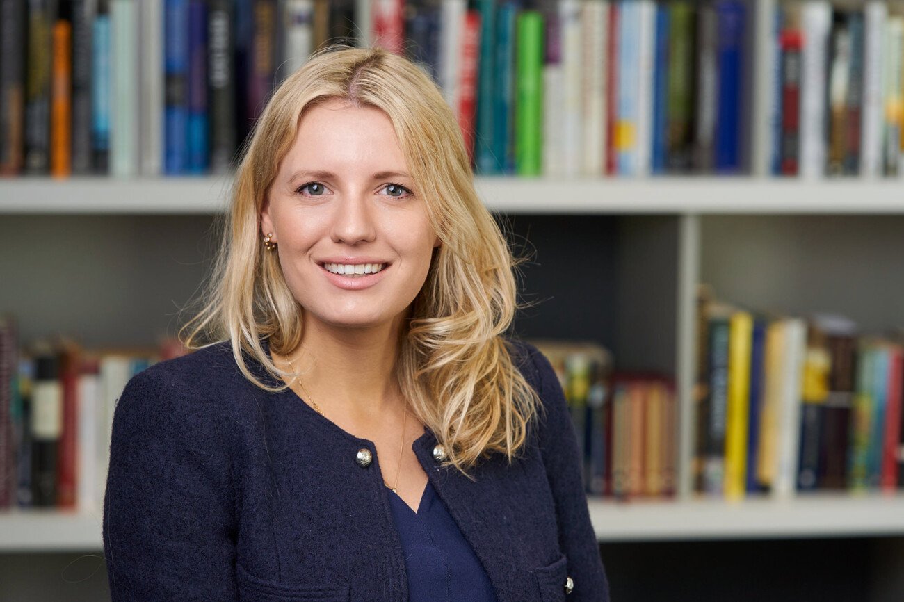 A corporate headshot photographer captures a person with long blonde hair, wearing a dark jacket and smiling in front of a bookshelf brimming with various books. James Gifford-Mead Photography - Event Photographer London