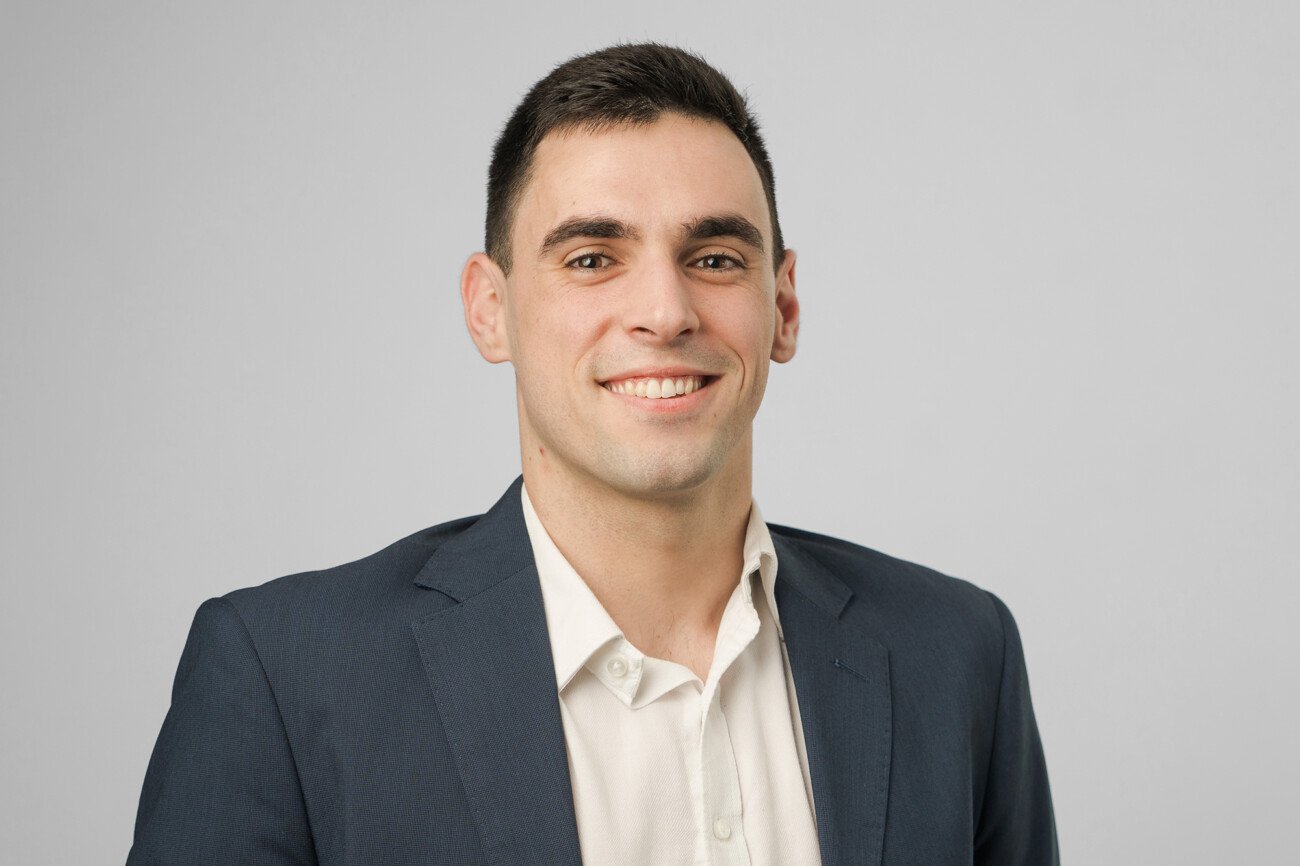 A person with short dark hair smiles confidently in a corporate headshot, wearing a dark suit and light-colored shirt against a plain, light gray backdrop. James Gifford-Mead Photography - Event Photographer London