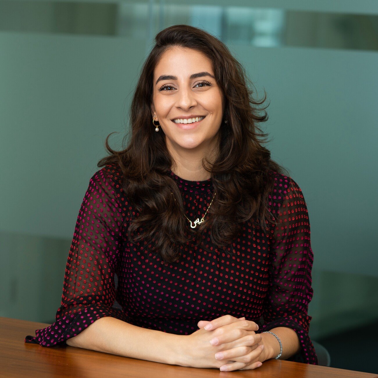 A woman with long dark hair sits at a wooden table, wearing a dark red patterned blouse. She smiles warmly, hands folded on the table, as if captured by a corporate headshot photographer. The background features frosted glass panels, adding a modern touch to the scene. James Gifford-Mead Photography - Event Photographer London
