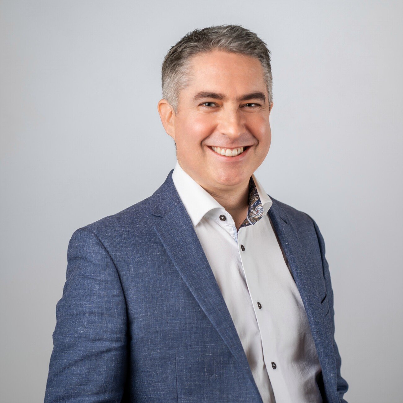 Smiling man in a blue blazer and white shirt, expertly captured by a corporate headshot photographer, stands against a plain light gray background. James Gifford-Mead Photography - Event Photographer London
