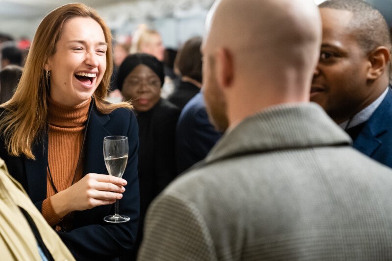 At a bustling London conference, a woman with long hair and a brown turtleneck laughs while holding a champagne glass. The photographer captures the lively atmosphere as several attendees in formal attire engage in animated conversation around her