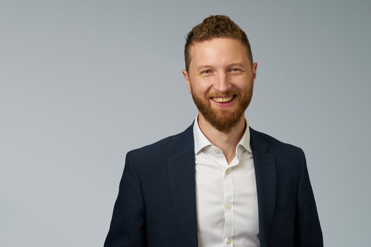 A man with curly hair and a beard smiles warmly at the camera, perfectly captured by a corporate headshot photographer. He is wearing a dark suit jacket over a white dress shirt against a light gray background. James Gifford-Mead Photography - Event Photographer London