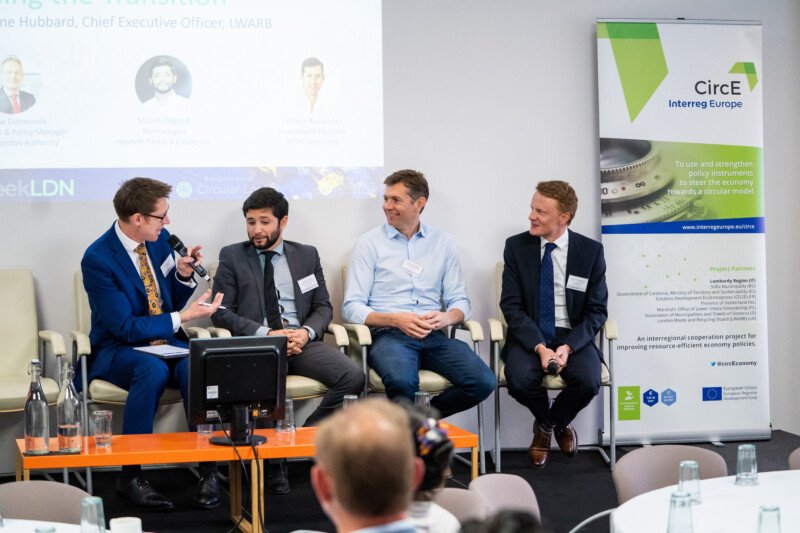 Four men in business attire are seated in a panel discussion at the London Conference. One speaks into a microphone while others listen attentively. Behind them is a screen and a CircE Interreg Europe roll-up banner, captured by a professional photographer. A table with glasses and a bottle is in the foreground