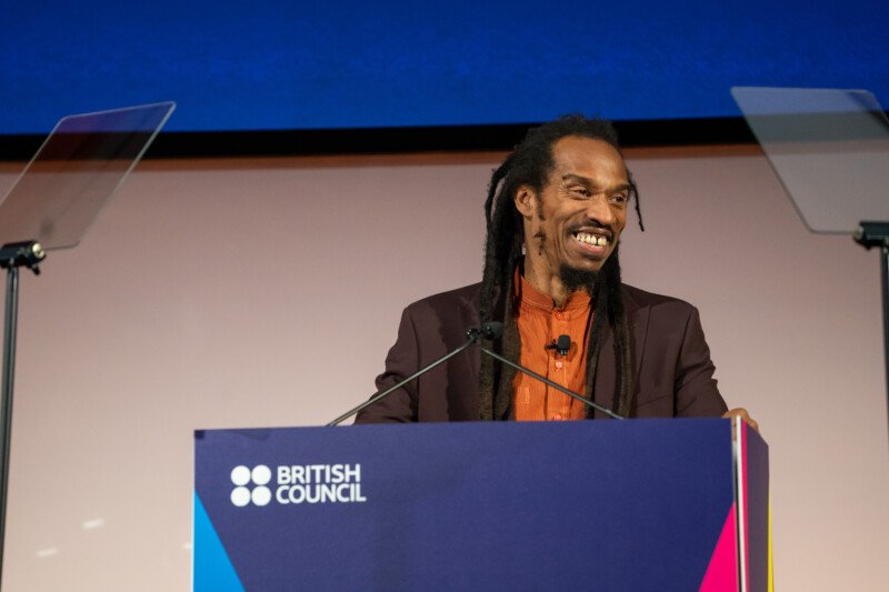 A man with long dreadlocks smiles at the podium, marked by the British Council logo. Captured by a London conference photographer, he's dressed in a dark blazer over an orange shirt, set against a blue and white backdrop with two transparent teleprompters on each side