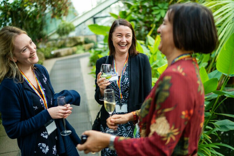 Three people smiling and holding drinks, engaged in conversation. They stand in a lush indoor garden with vibrant green plants, perfectly captured by a London conference photographer. Two are wearing lanyards, while one is holding a wine glass