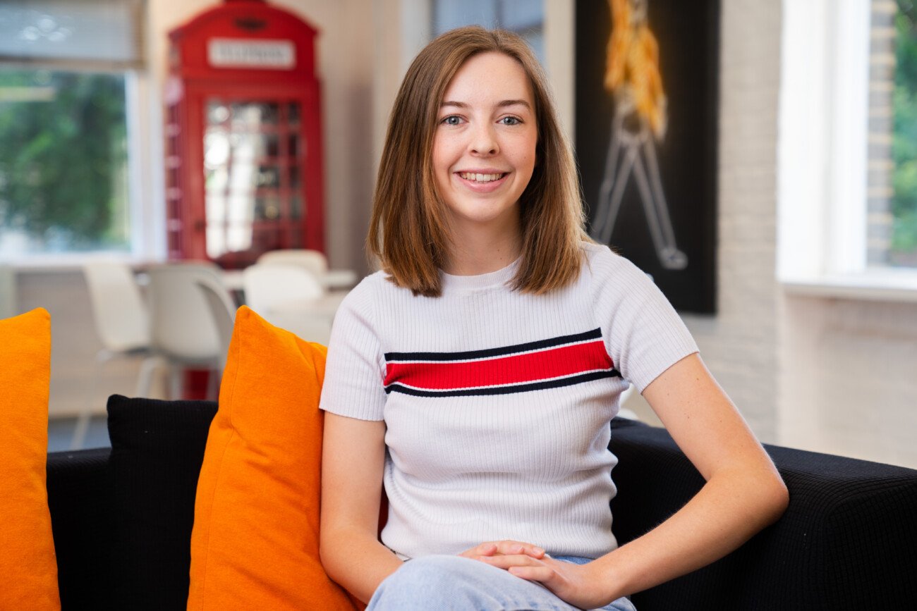A woman with shoulder-length hair sits on a black couch with orange cushions, smiling confidently at the camera. Her white top features red and black stripes. In the background, there's a red telephone booth and modern decor, perfectly captured by the skilled corporate headshot photographer. James Gifford-Mead Photography - Event Photographer London
