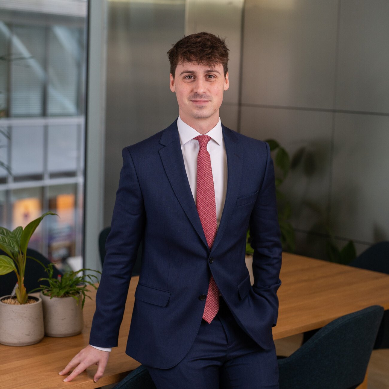 A person in a navy suit and red tie poses for a corporate headshot beside a wooden table adorned with potted plants. The glass wall backdrop showcases reflections and modern architecture elements, capturing a perfect blend of professionalism and style. James Gifford-Mead Photography - Event Photographer London