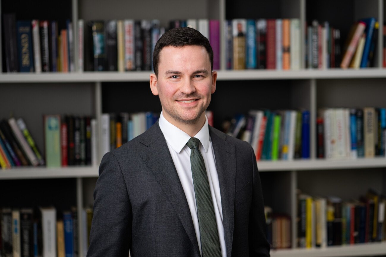 A smiling man in a gray suit and green tie stands in front of a bookshelf filled with books, perfectly captured by the corporate headshot photographer. James Gifford-Mead Photography - Event Photographer London