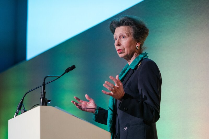 An older woman with short gray hair and a green scarf speaks at a podium, microphones poised to capture every word. The backdrop is a gradient of green and blue, reminiscent of the London Conference. She gestures animatedly, embodying the finesse of an experienced photographer