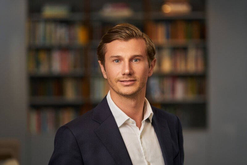A person with short, light brown hair is wearing a dark suit and white shirt, standing in front of a blurry background of bookshelves. Captured by a skilled corporate headshot photographer, the expression is neutral yet professional. James Gifford-Mead Photography - Event Photographer London