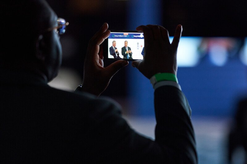 A London Conference photographer captures a photo of two individuals speaking on stage. The background is dark, and the screen displays the speakers clearly, their gestures visible. The photographer holding the smartphone is silhouetted against the scene