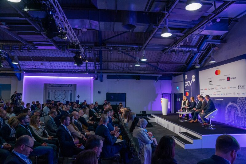 A large audience in a modern conference room listens to a panel of five speakers on stage, captured by a talented London conference photographer. The setting features purple lighting and banners with logos, while attendees are seated in rows under an industrial ceiling design