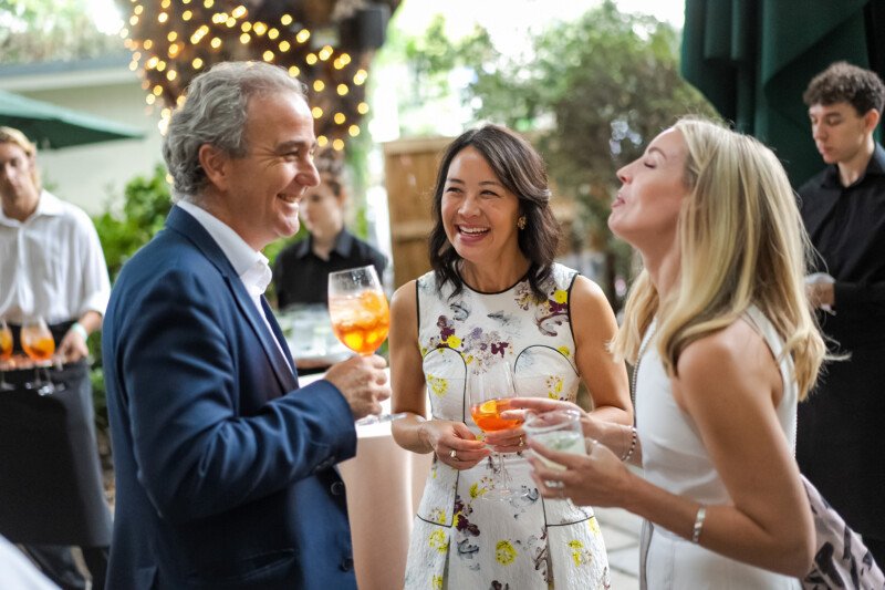 Three people in formal attire laugh and talk while holding drinks at an outdoor gathering. Captured by a skilled London conference photographer, string lights and trees create a festive atmosphere, with others in the background contributing to the lively setting