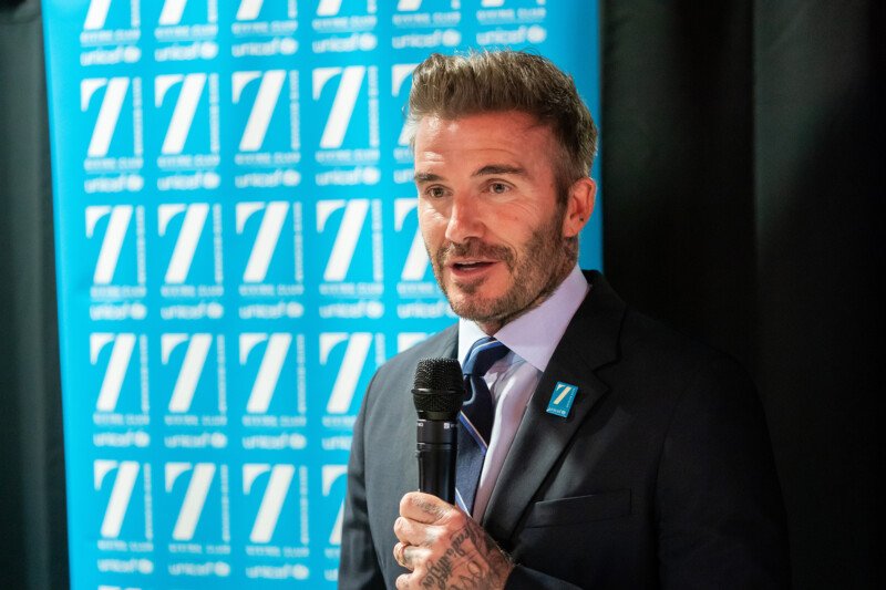 At a London conference, a man with short hair and a beard speaks into the microphone. Dressed in a dark suit and light blue tie, he stands confidently against a backdrop adorned with a repeated blue and white logo pattern, perfectly captured by the photographer