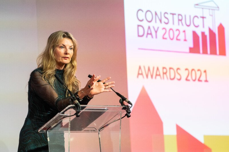 A woman with long blonde hair stands at a clear podium, speaking at a formal event, the "Construct Day 2021 Awards," captured by a skilled London Conference Photographer. Her dark outfit complements the professional ambiance