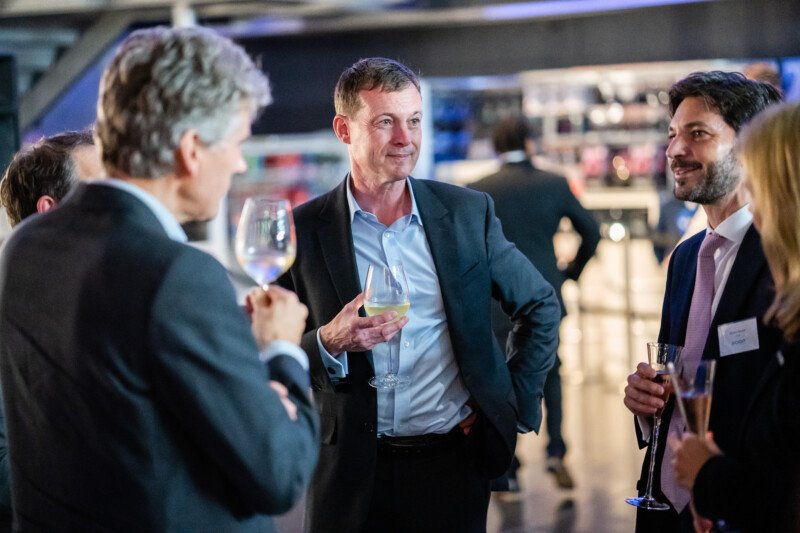 At a London conference, a group in formal attire mingles while a man at the center holds a wine glass. The photographer captures guests around him, all engaged in animated conversation, their drinks held aloft amidst the softly blurred backdrop of this elegant social gathering