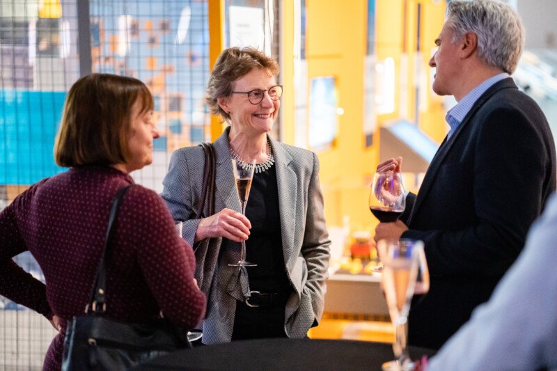 At a lively social gathering, three people converse with wine glasses in hand, framed by bright and colorful decor. Capturing this vibrant moment is akin to a London conference photographer's keen eye for atmospheric and engaging scenes