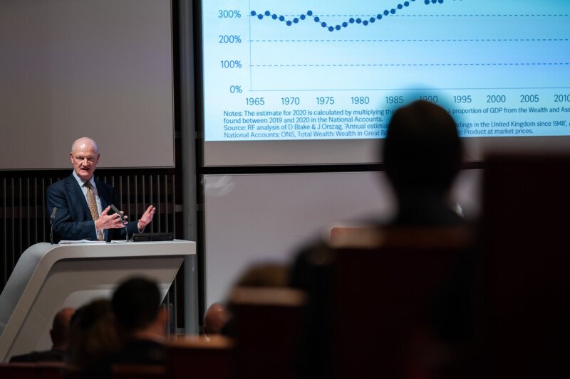 A speaker at a podium addresses an audience in a lecture hall, captured by a London conference photographer. A large screen displays a graph showing data trends from 1965 to 2020, with a noticeable upward trajectory