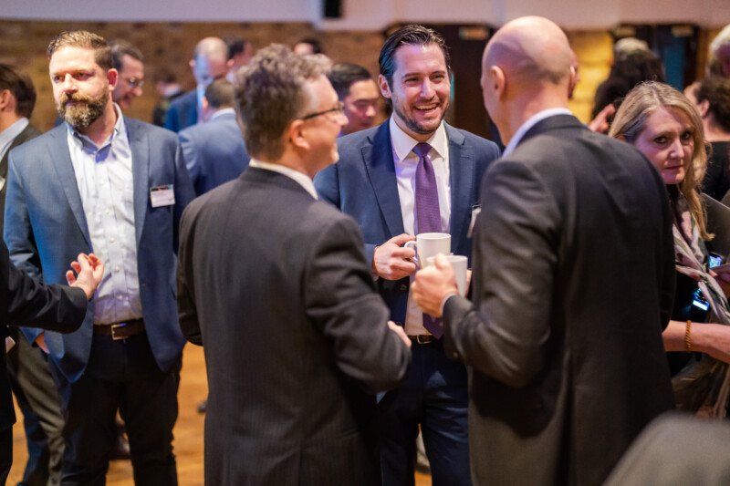 A group of people in business attire are standing and conversing at a London conference. One man in the center holds a white mug, smiling. The background shows more attendees engaged in discussions, capturing the vibrant networking atmosphere through the lens of a skilled photographer