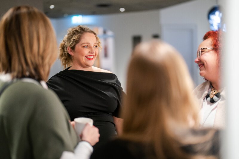 A group of four are engaged in a lively conversation indoors, perhaps at a London conference. One person in a black outfit, possibly the event photographer, is smiling and listening attentively. The setting has the relaxed yet purposeful vibe of a casual gathering or meeting