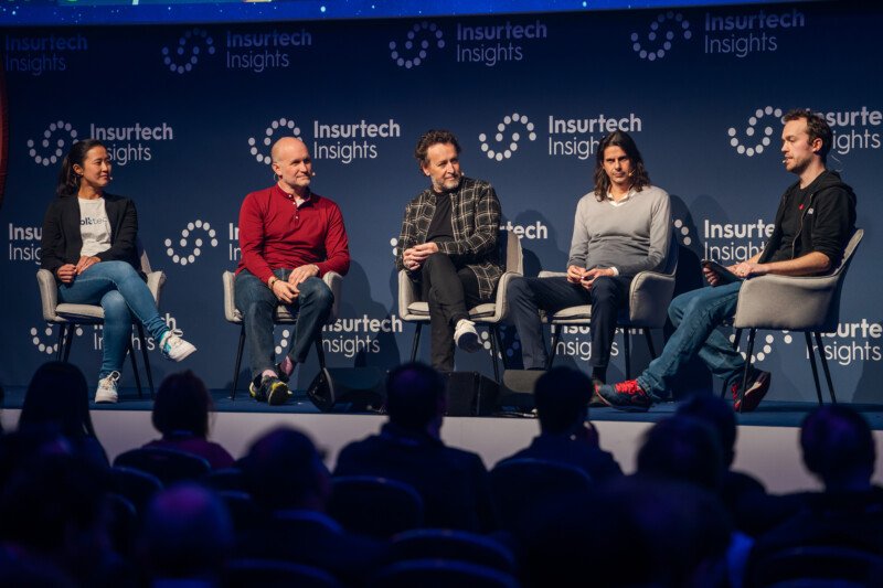 Five people sit in chairs on a stage during a panel discussion at an Insurtech Insights event, expertly captured by a London conference photographer. The backdrop features the event's logo, while an attentive audience fills the foreground, all eyes on the insightful panelists