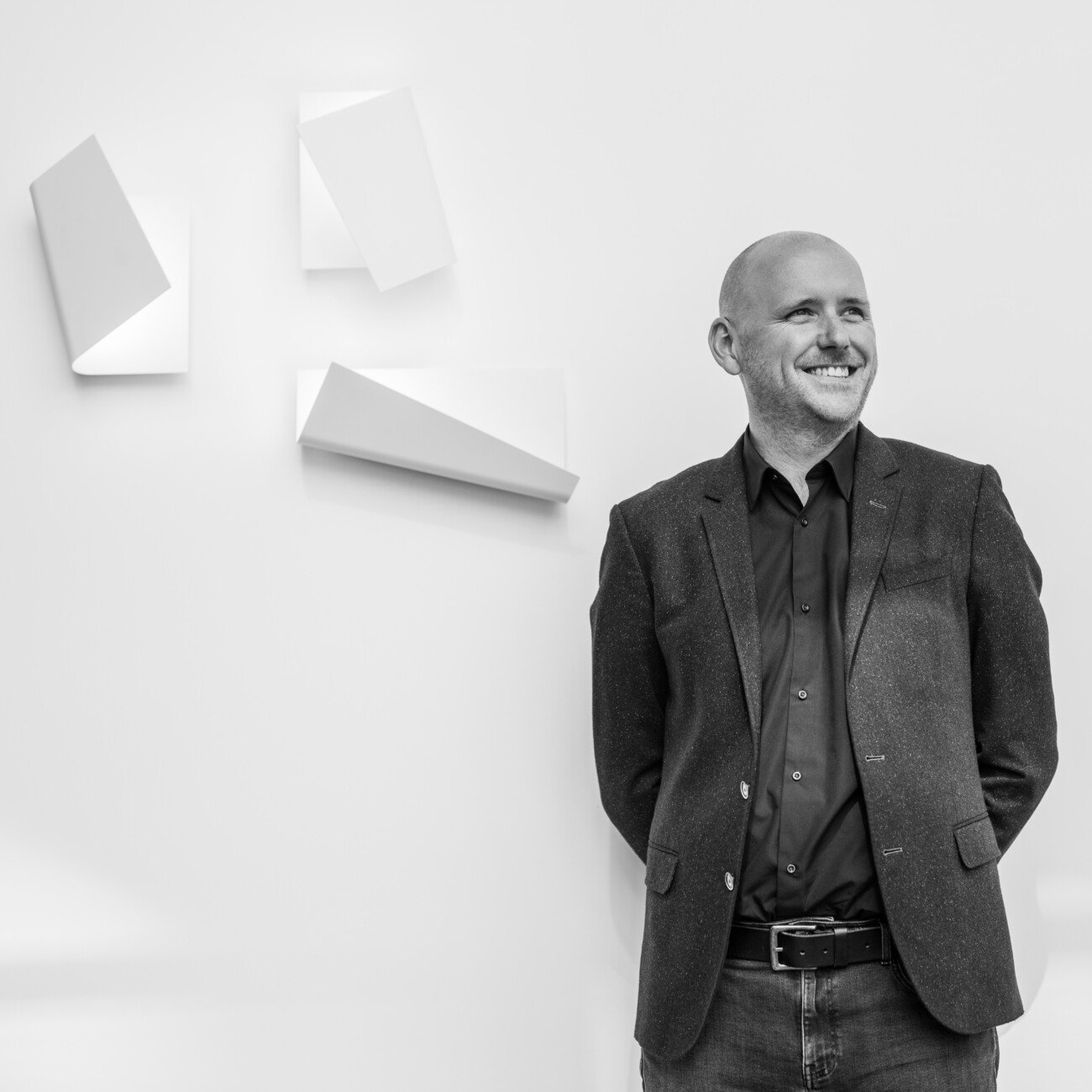 A man stands smiling with his hands behind his back, wearing a dark blazer and jeans. Captured by a corporate headshot photographer, he is positioned against a plain white wall adorned with three abstract white shapes. The image is in black and white. James Gifford-Mead Photography - Event Photographer London