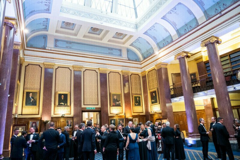 A formal gathering in an elegant hall with high ceilings, ornate decorations, and framed portraits reminiscent of a London conference. Attendees in formal attire mingle and converse under soft lighting as a photographer discreetly captures the moment. A balcony with bookshelves overlooks the scene