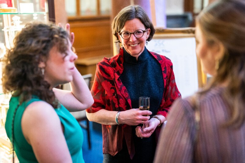 At a London conference, a woman in a red patterned jacket holds a glass and smiles while conversing with two other women. The softly lit background creates a warm and inviting atmosphere, perfectly captured by the talented photographer