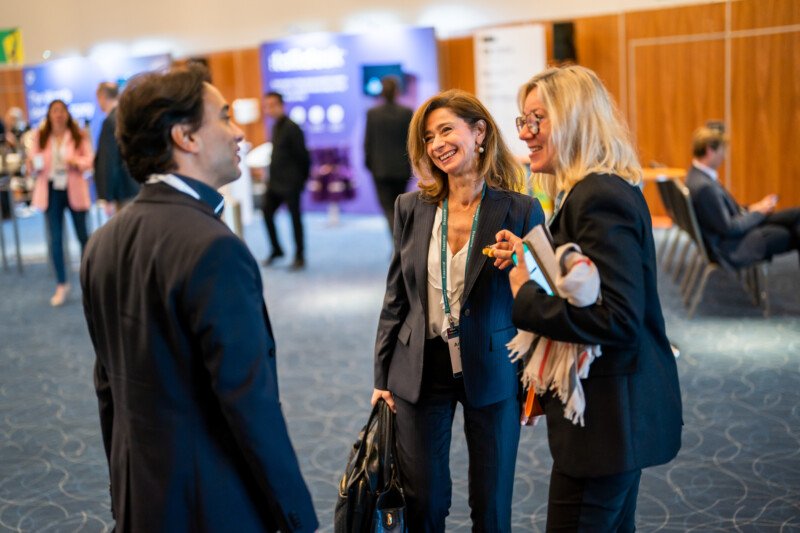 Amidst the vibrant scene of a London conference, a photographer captures three professionals engaged in lively conversation. Two women, lanyards and bags in hand, share smiles with a suited gentleman against a bustling backdrop of booths and attendees