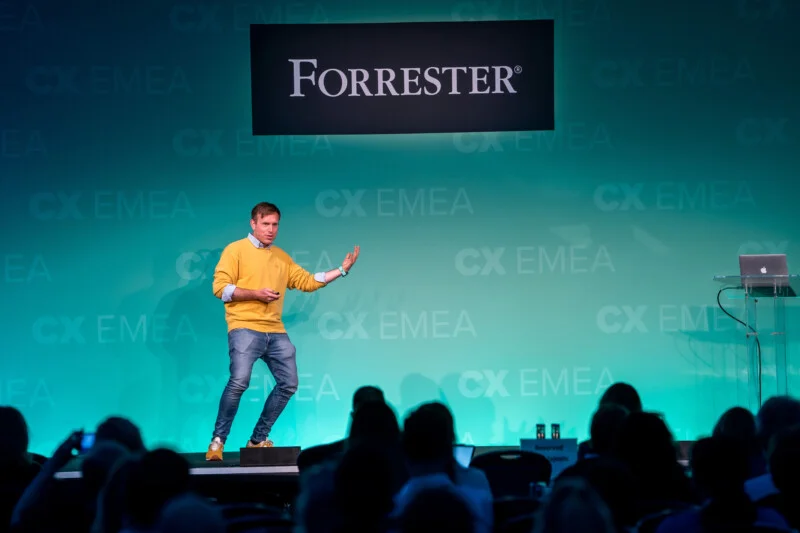 A speaker in a yellow sweater and jeans gestures enthusiastically on stage at the Forrester CX EMEA event. Captured by a London conference photographer, the audience is visible in the foreground, set against a turquoise backdrop with the event's logo shining brightly behind