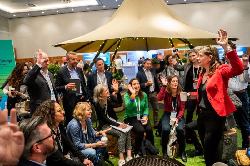 In a conference room beneath a large canopy, a London photographer captures the dynamic scene of people seated and standing, some with raised hands. They are actively engaging in the discussion led by a woman in a red jacket