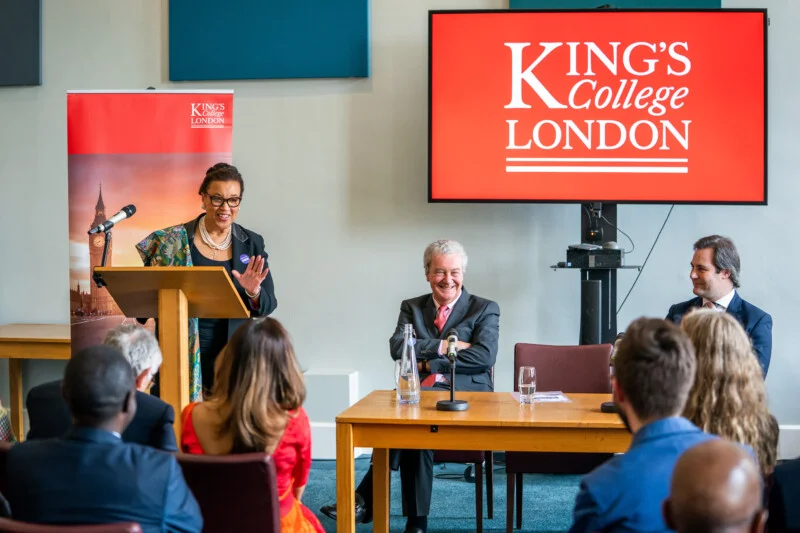 A photographer captures a speaker at a podium addressing an audience at King's College London's conference. A large screen displays the college’s name, while two people are seated at a table on stage, engaging with the attendees