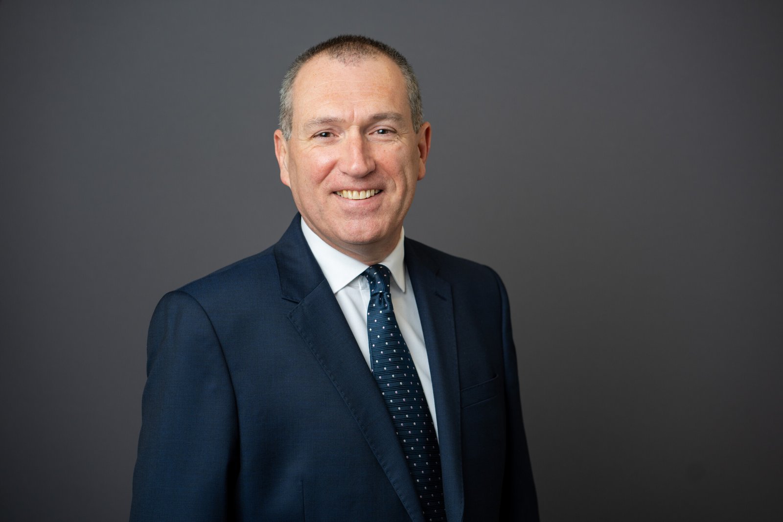 A man in a dark suit and polka dot tie smiles confidently at the camera. His short hair complements the professional aura captured by a skilled corporate headshot photographer, all set against a plain, dark background