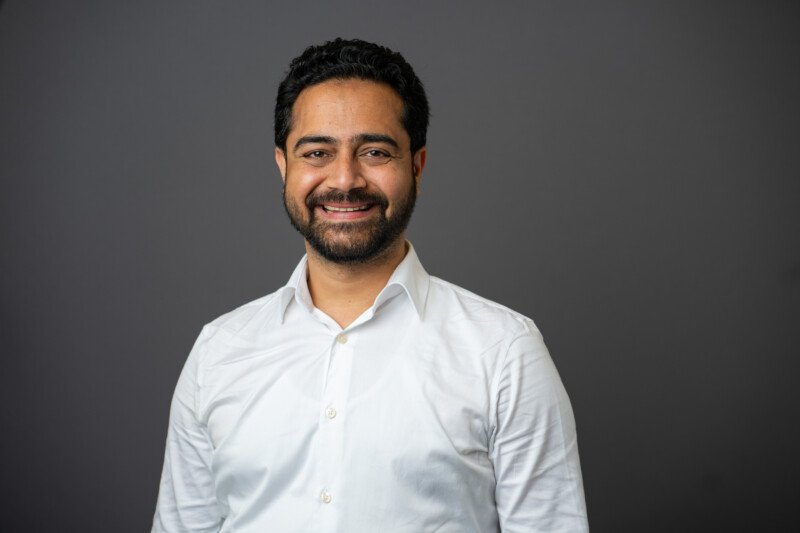 A smiling man with a beard dons a white button-up shirt against a plain gray background, perfectly captured by a skilled corporate headshot photographer. James Gifford-Mead Photography - Event Photographer London