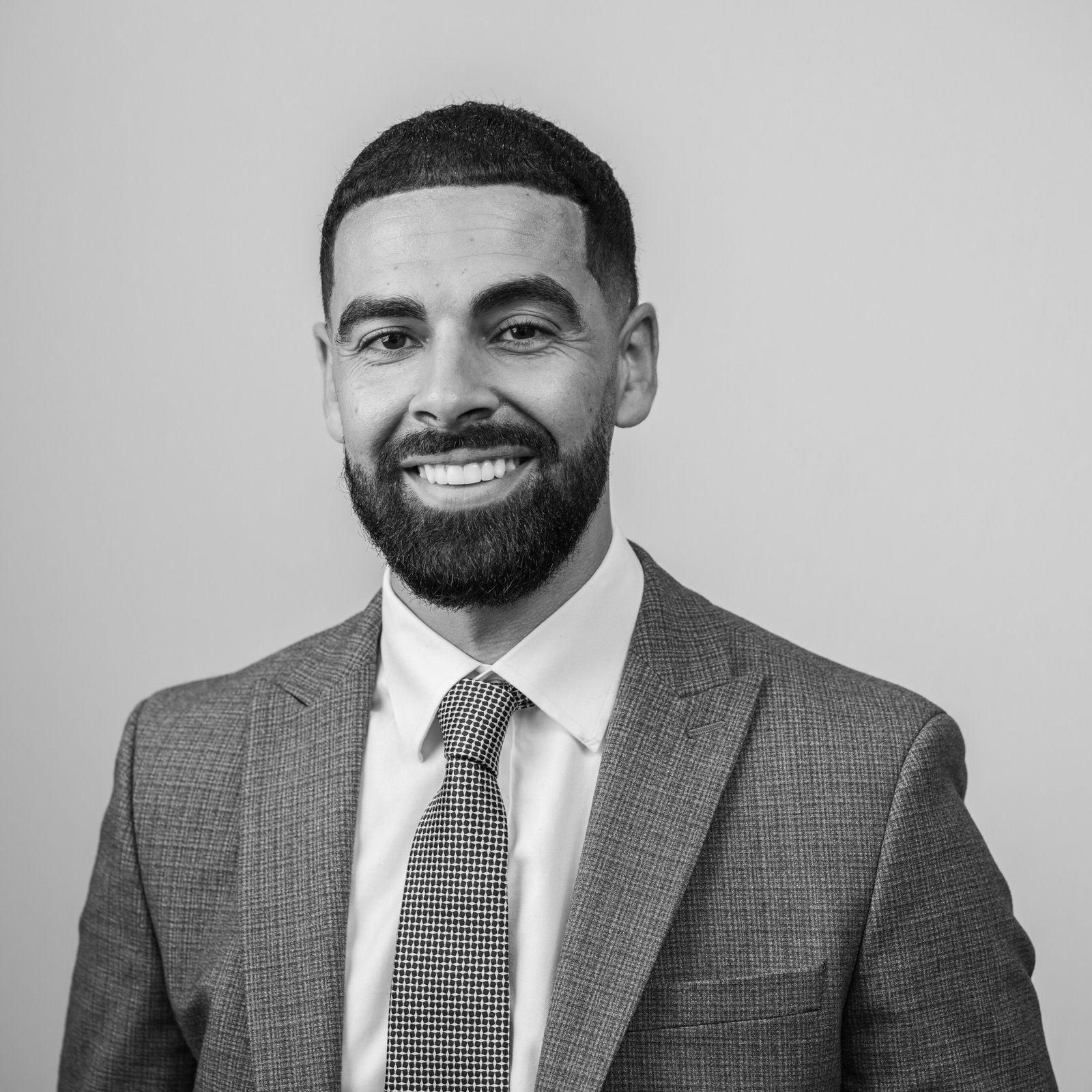 Black and white portrait of a smiling man with a beard, styled by a corporate headshot photographer. He wears a suit jacket, white shirt, and patterned tie, facing the camera against a plain backdrop