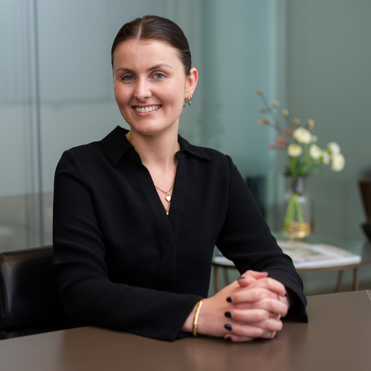A woman with dark hair, clad in a black outfit and gold jewelry, sits at a table with her hands folded. She is smiling in an office space adorned with flowers—a perfect capture by a corporate headshot photographer. James Gifford-Mead Photography - Event Photographer London