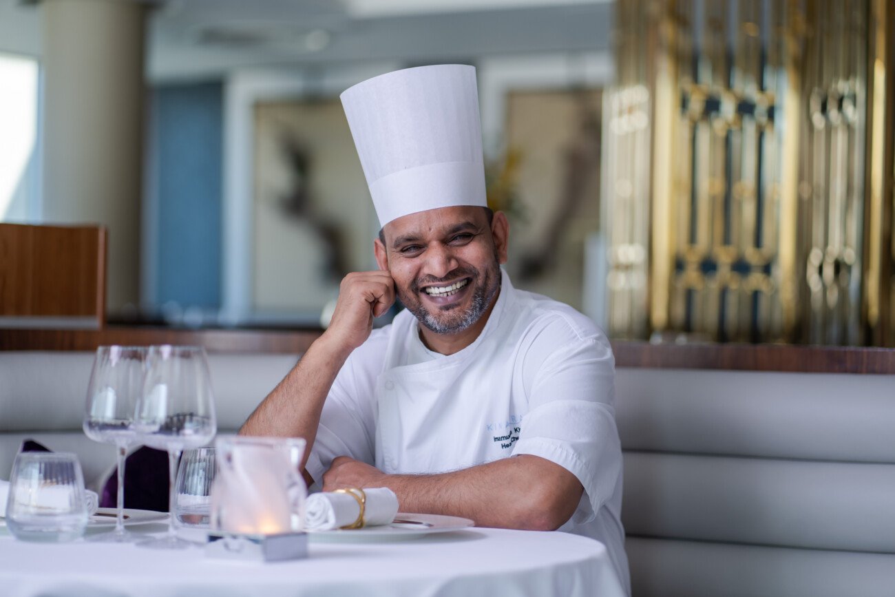 A chef in a white uniform and tall hat sits at a restaurant table, smiling with his hand on his face, as if posing for a corporate headshot photographer. The table is adorned with wine glasses, a candle, and a rolled napkin against an elegantly decorated background. James Gifford-Mead Photography - Event Photographer London