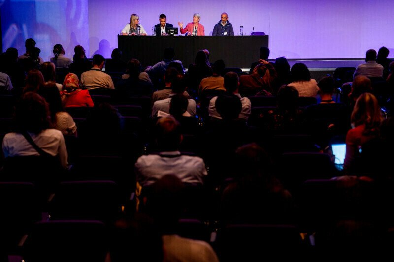 At a bustling London conference, a panel of four speakers sits at a long table on stage against a vivid purple backdrop. The audience listens intently, with several attendees taking notes as the conference photographer captures these engaging moments