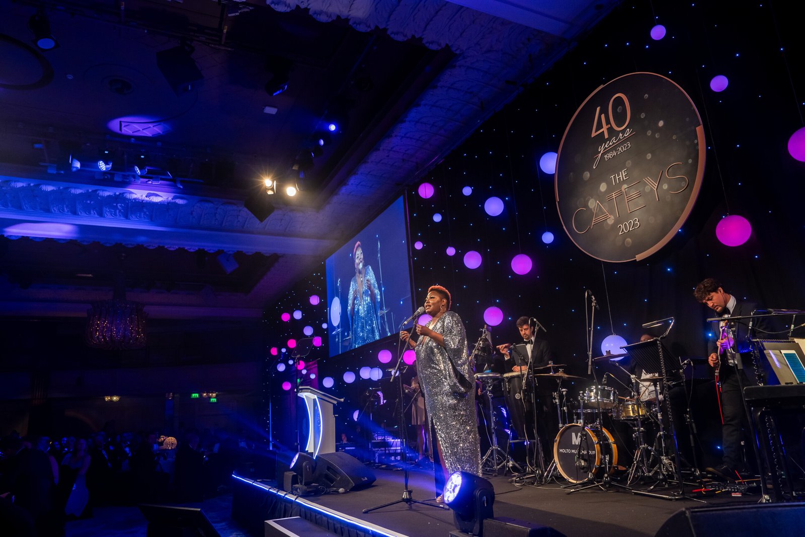 A performer in a sparkly outfit sings on stage at a gala event, where an awards photographer captures the moment. The stage is adorned with round lights and a sign reading "40 Awards, The CA News 2023," complemented by a band and a screen displaying the vibrant scene