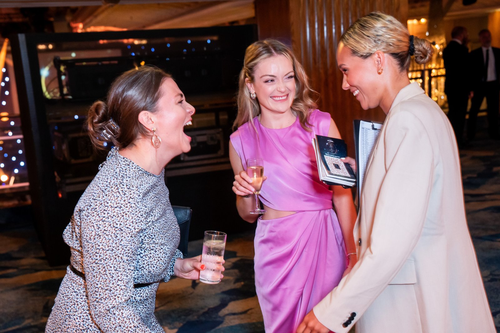 Three women joyfully converse at an indoor event, captured perfectly by the awards photographer. One wears a patterned dress, another a pink dress holding a drink, and the third in a light blazer with a booklet. They're smiling and laughing together in the warmly lit setting. James Gifford-Mead Photography - Event Photographer London