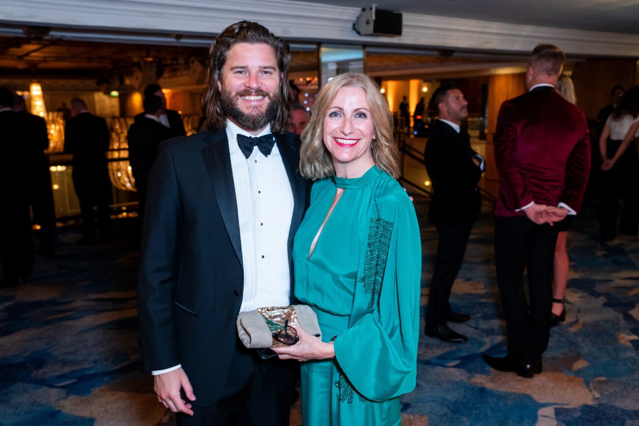 A man in a black tuxedo and a woman in a teal dress pose together at an event, captured by an awards photographer. The woman holds a small clutch as people in formal attire mingle in the background