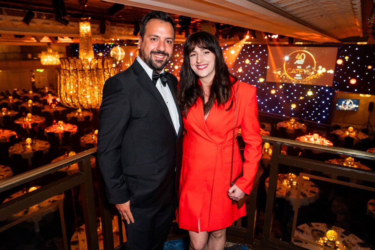 A man in a tuxedo and a woman in a red dress smile at the camera, captured by an awards photographer. They stand on a balcony overlooking a glamorous event, where chandeliers sparkle and a large screen prominently displays "40 Years" in the background