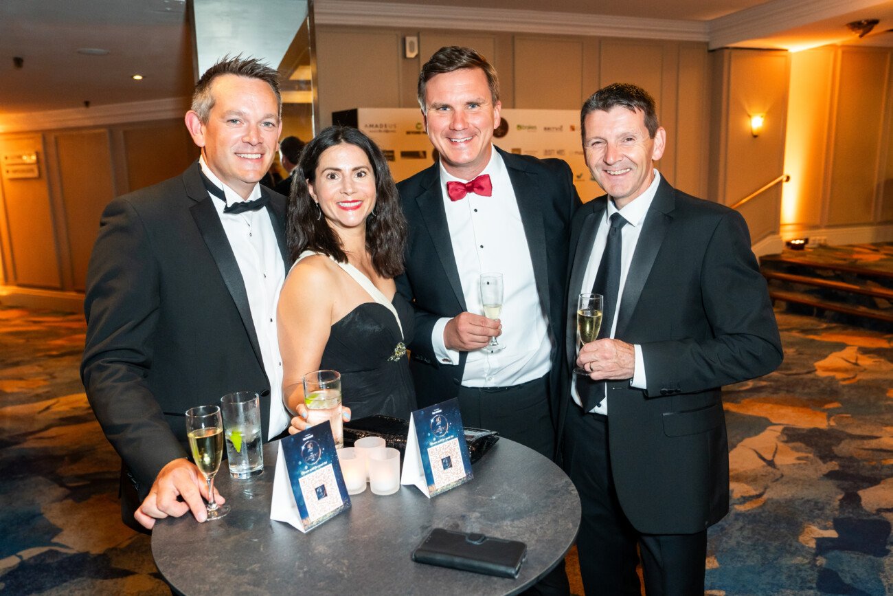 Four people in formal attire stand around a table, smiling for the awards photographer. Three men in tuxedos and a woman in a black dress hold glasses of champagne. The table is adorned with candles and small awards, set against the elegant backdrop of a carpeted hall