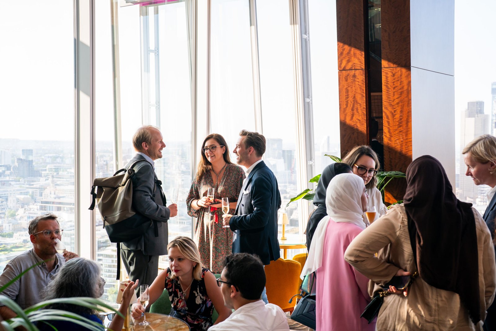 A group of people socializing in a modern lounge with large windows offering a city view. Some are standing with drinks, others sitting or talking in small groups. Diverse clothing and casual atmosphere suggest a relaxed gathering or event