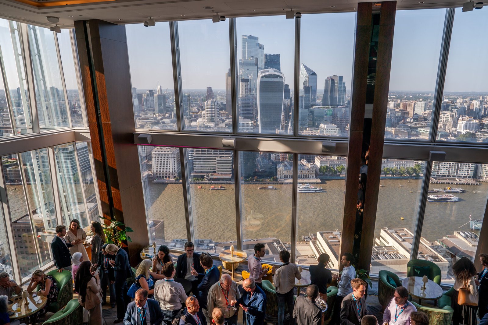A group of people socializing in a glass-walled room with a panoramic view of a cityscape, featuring tall buildings and a river. Sunlight filters through the windows, creating a bright and lively atmosphere