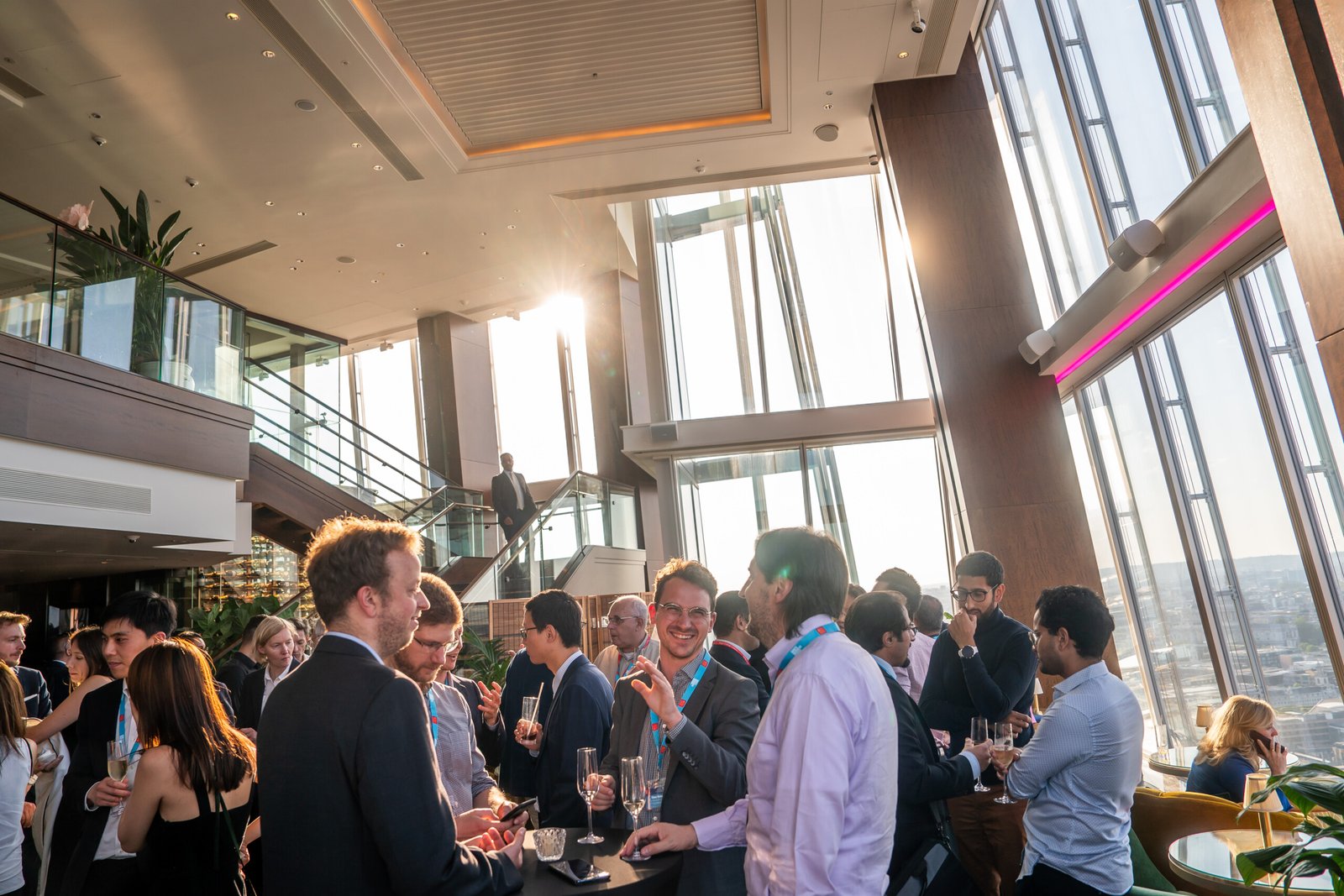 People gathered at a modern rooftop event with tall windows and a city view. They are socializing and holding drinks, while sunlight streams in through the glass. The atmosphere is lively and upscale