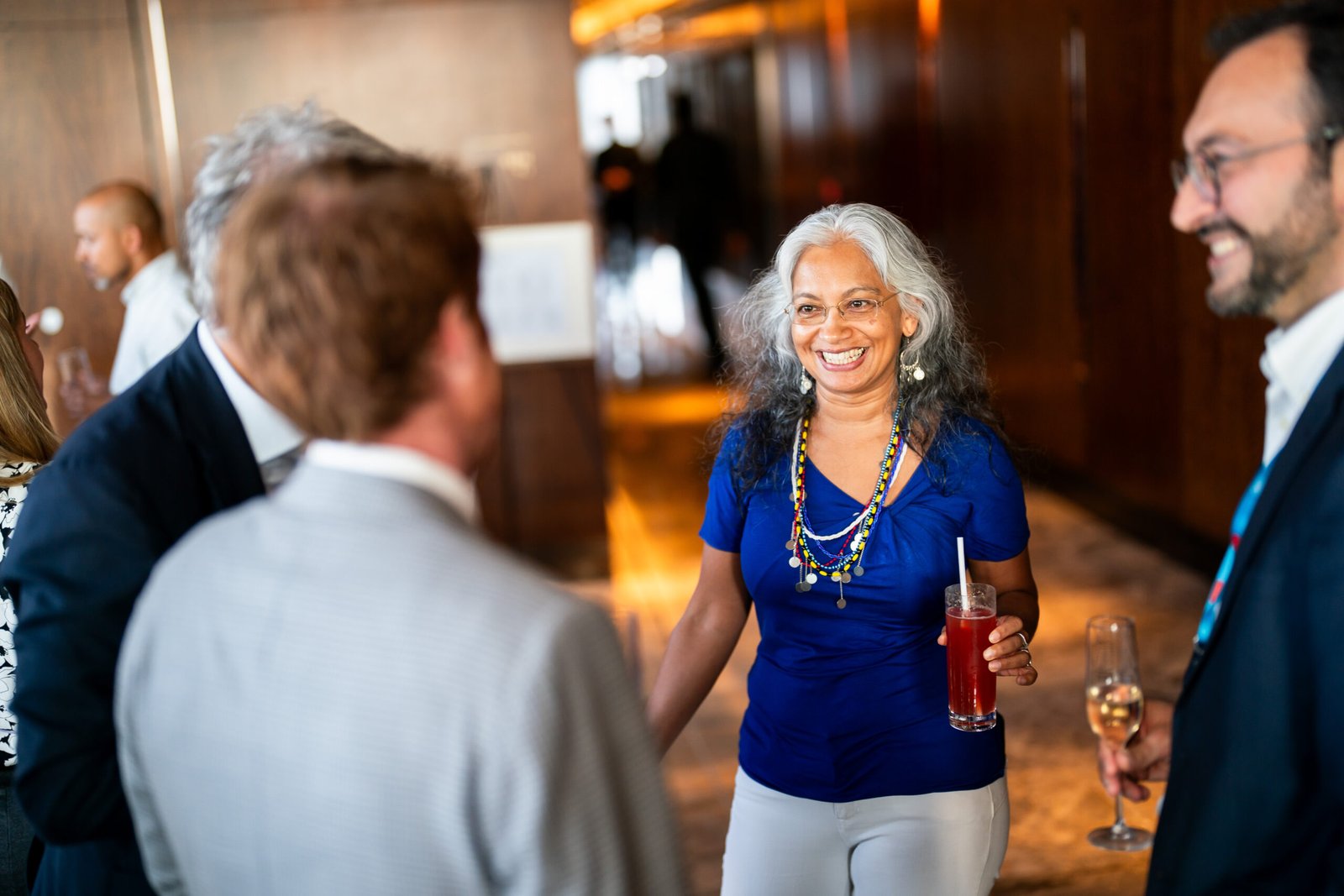 A woman with long gray hair, wearing a blue top and necklaces, smiles while holding a red drink. She's engaged in conversation with people in a bright indoor setting. Other individuals in the background are also holding drinks