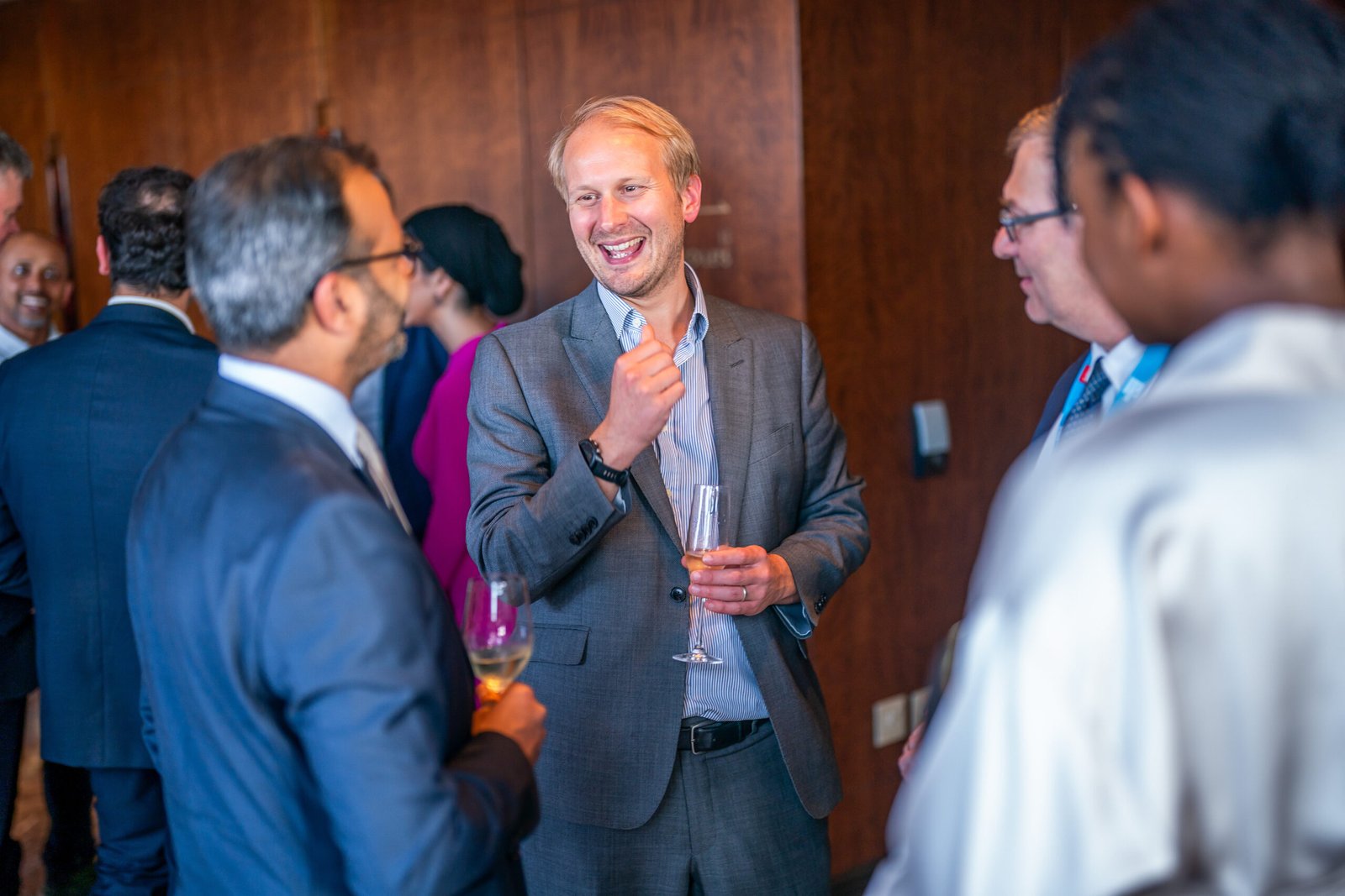 A group of people in formal attire engage in conversation at a social event. A man in a gray suit, smiling, holds a glass and gestures with his hand. The background is blurred, focusing on the group
